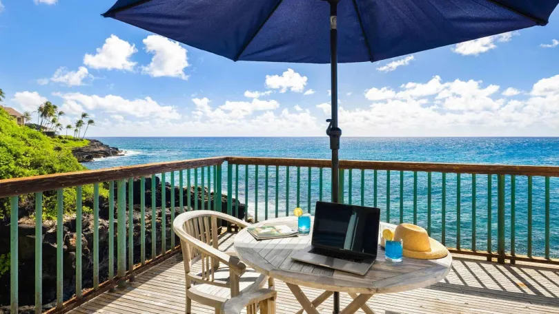 Ein Tisch mit Laptop und Stuhl unter einem Sonnenschirm auf einer Terrasse mit Meeresblick.