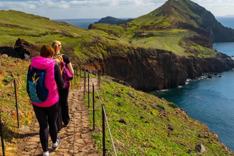 Drei Personen wandern auf einem schmalen, gepflasterten Pfad entlang einer Klippe mit Blick auf das Meer.