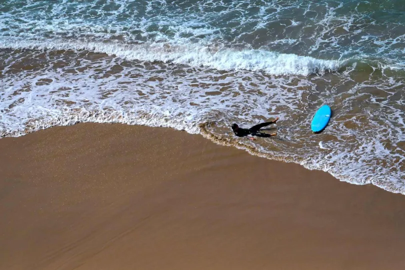 ine Person im Neoprenanzug liegt am Strand neben einem Surfbrett.