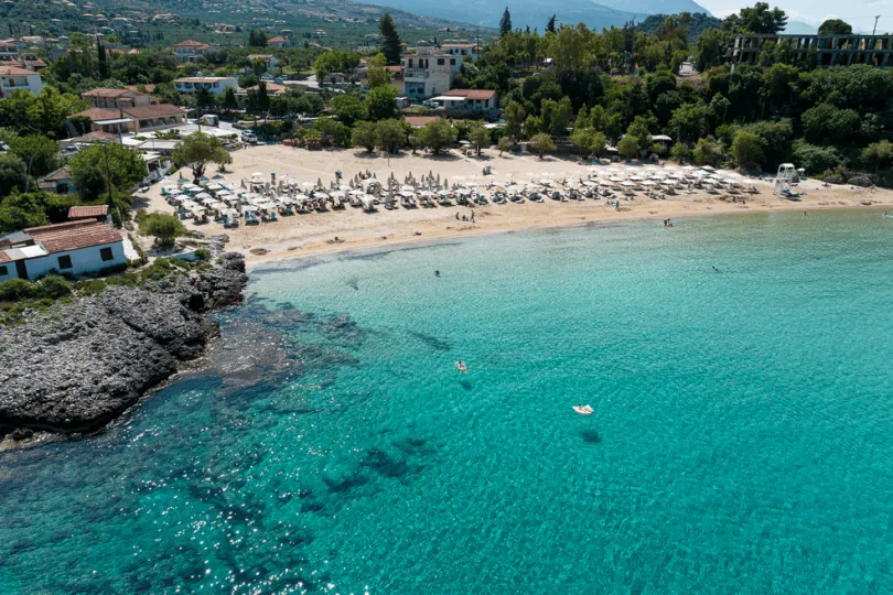 Strand mit Liegen, türkisfarbenem Wasser und umliegenden Gebäuden im Grünen.