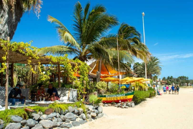 Strandrestaurant „Barracuda“, Palmen, sonniger Tag, Gäste am Strand.