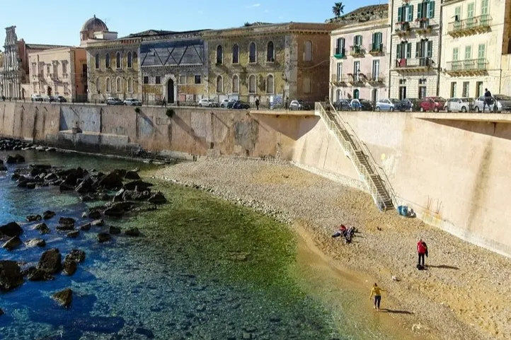 felsige Küste mit Strand, Treppe, alten Gebäuden und geparkten Autos.