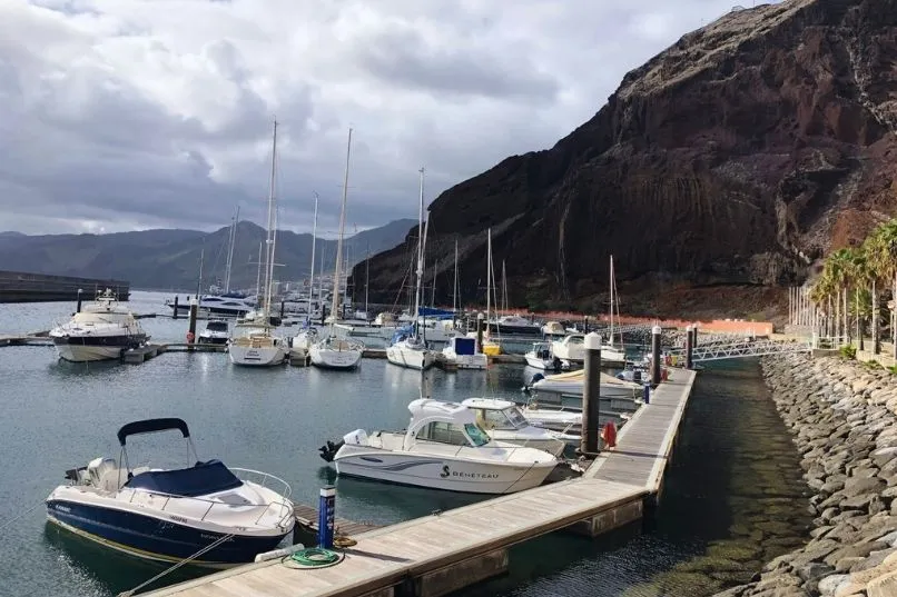 Boote liegen im ruhigen Hafen vor einer steilen Felswand unter wolkigem Himmel.