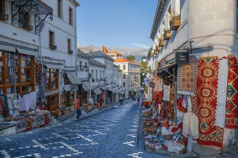  Gjirokastra – charmante Altstadtgasse mit Kopfsteinpflaster, weißen Häusern und traditionellen Teppichen.