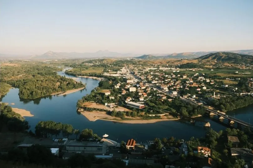 Panoramablick auf eine albanische Stadt am Fluss, umgeben von Hügeln und grüner Landschaft.