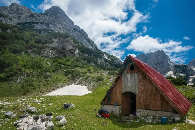 Almhütte mit rotem Dach in den montenegrinischen Bergen, umgeben von grüner Natur und felsigen Gipfeln.
