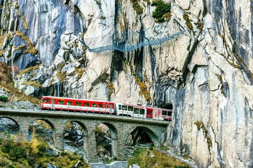 Roter Zug fährt über ein steinernes Viadukt durch dramatische Felslandschaft in den Alpen.