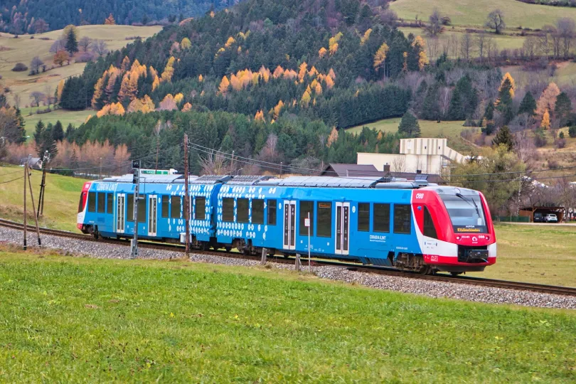 Ein moderner blau-roter Regionalzug fährt durch eine grüne Hügellandschaft mit herbstlich gefärbten Bäumen im Hintergrund.