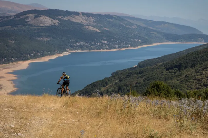 Mountainbiker fährt auf einem Hügel oberhalb des Mavrovo-Sees in Nordmazedonien mit Blick auf das weite Wasser und die bewaldeten Berge im Hintergrund.