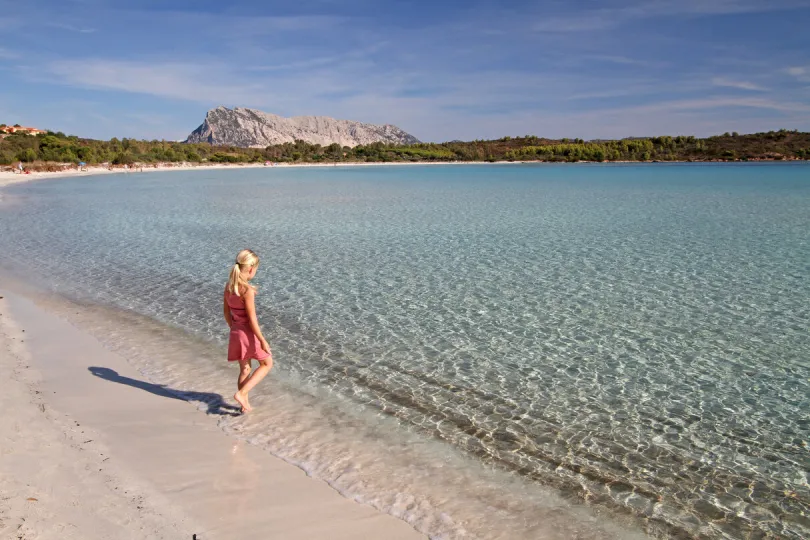 Ein weitläufiger Strand auf sardinien mit türkis blauem Wasser und einem Kind das im seichten Wasser steht.
