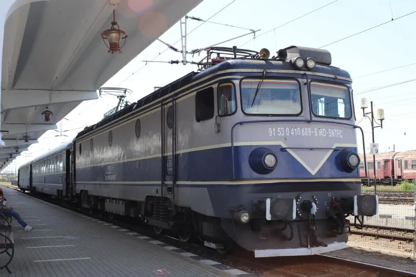 Eine blau-graue Elektrolokomotive steht mit mehreren Waggons an einem Bahnsteig unter einem überdachten Gleisbereich.