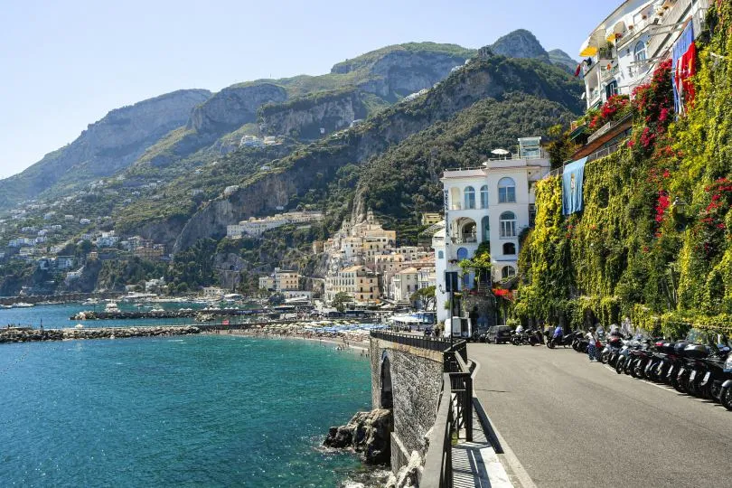 Eine Küstenstraße mit parkenden Motorrollern verläuft entlang eines türkisblauen Meeres, vorbei an mit Pflanzen bewachsenen Häusern, während sich eine malerische Stadt an steilen Berghängen im Hintergrund erstreckt.