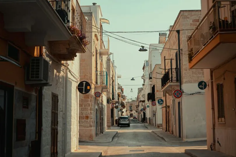 ine schmale, ruhige Straße in einer mediterranen Altstadt, gesäumt von hellen Häusern mit Balkonen, Fensterläden und Verkehrszeichen, mit einem parkenden Auto im Hintergrund.