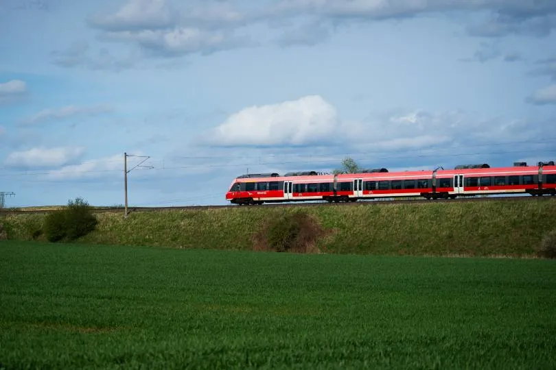 Roter Regionalzug der Deutschen Bahn fährt auf einer Bahnstrecke durch eine grüne Landschaft unter bewölktem Himmel.