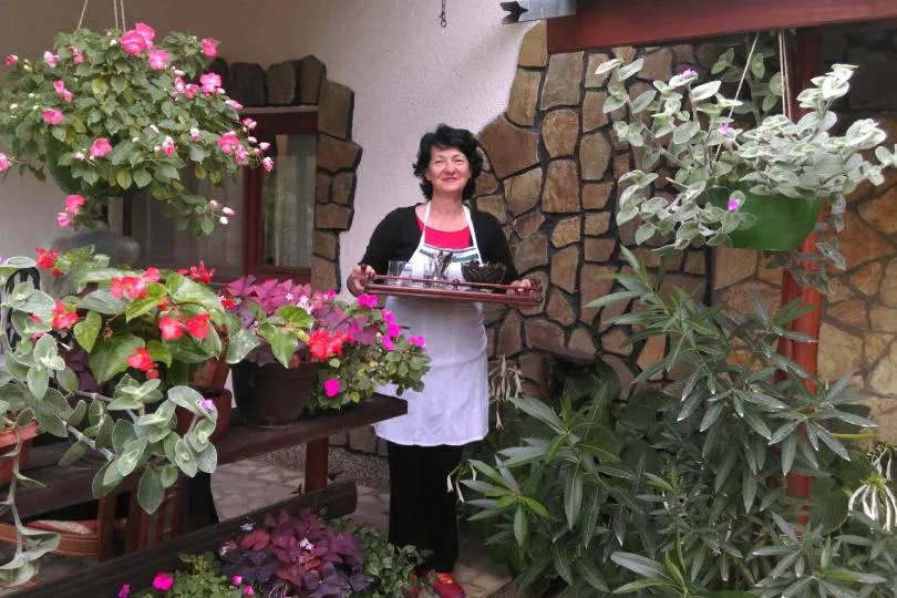 Frau mit Schürze steht auf einer Terrasse zwischen vielen blühenden Topfpflanzen und hält ein Tablett mit Getränken vor einem Haus mit Steinwand.