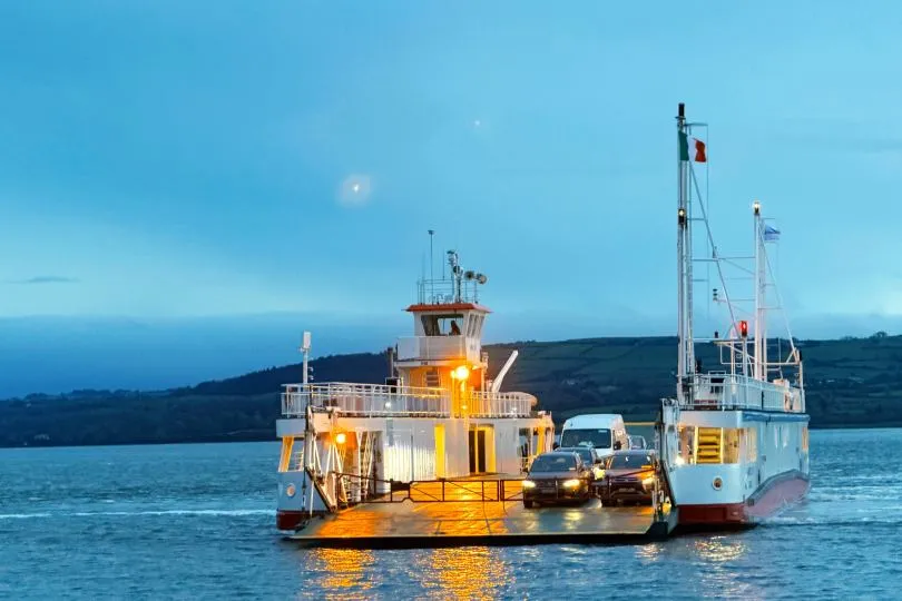 Fähre mit Autos an Bord überquert bei Dämmerung ein ruhiges Gewässer, Lichter spiegeln sich auf der Wasseroberfläche.