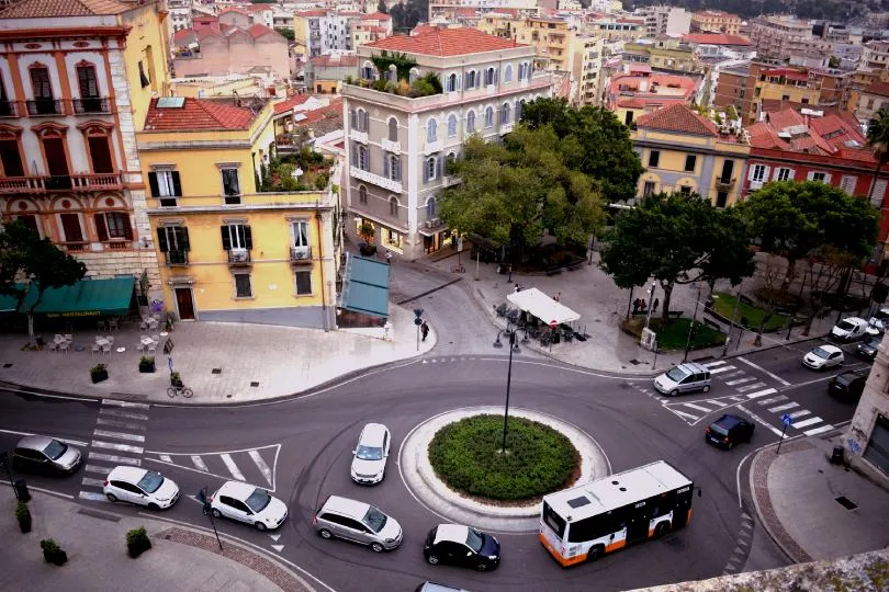 Ein Kreisverkehr in einer südeuropäischen Stadt, umgeben von mehrstöckigen Gebäuden mit roten Ziegeldächern, während Autos und ein Bus den Verkehr passieren und Menschen sich auf einem kleinen Platz mit Bäumen aufhalten.