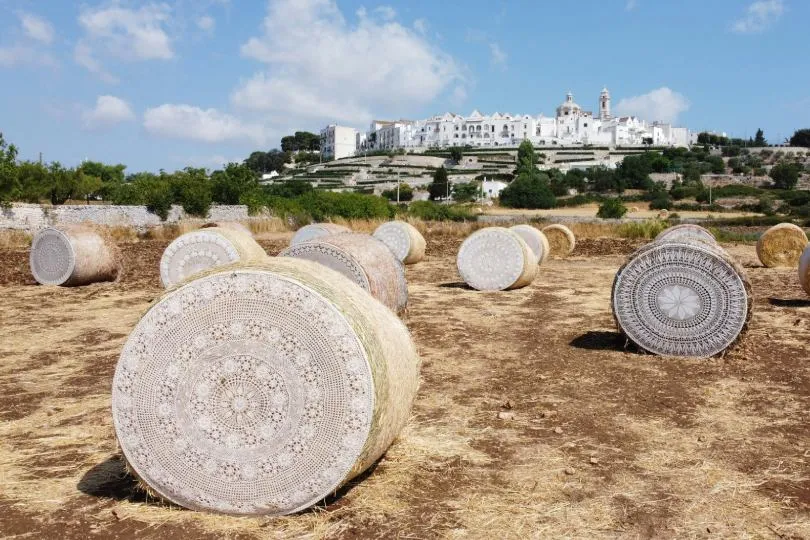 Heuballen auf einem Feld vor einer weißen Hügelsiedlung mit Kirche unter blauem Himmel in Apulien, Italien