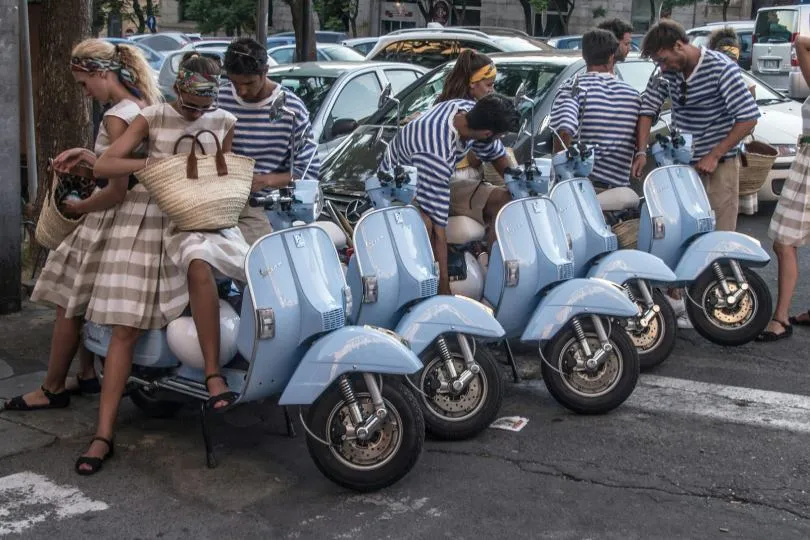 Gruppe von Menschen steht neben mehreren hellblauen Motorrollern in einer Stadtstraße und bereitet sich auf die Fahrt vor.