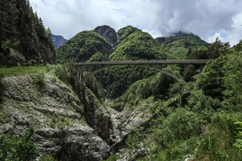 Bogenbrücke spannt sich über eine tiefe, felsige Schlucht in einer grünen Berglandschaft mit bewaldeten Hängen und bewölktem Himmel.