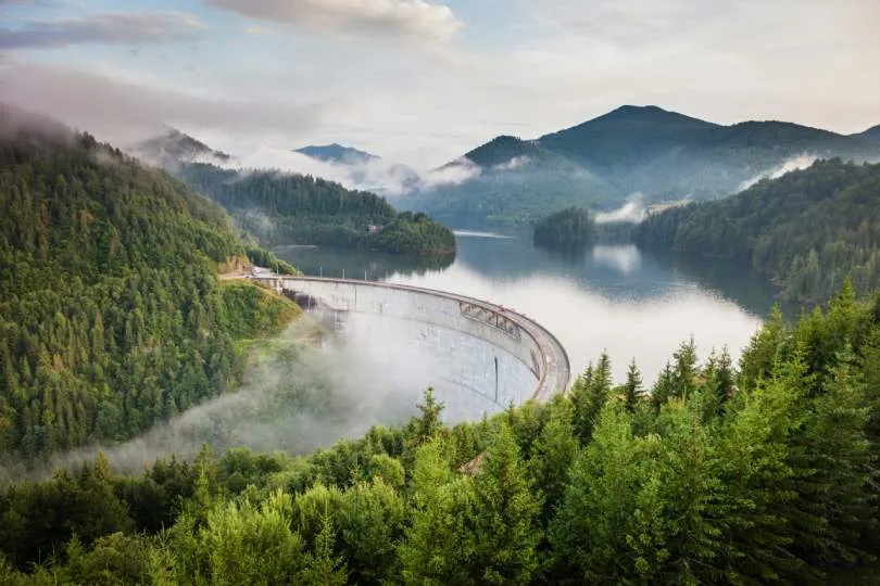 Ein großer, gebogener Staudamm liegt eingebettet in einer bewaldeten Berglandschaft. Hinter dem Damm erstreckt sich ein ruhiger See, umgeben von dichten Wäldern und sanften Hügeln. Nebelschwaden ziehen durch die Täler und verleihen der Szene eine ruhige, fast mystische Atmosphäre.