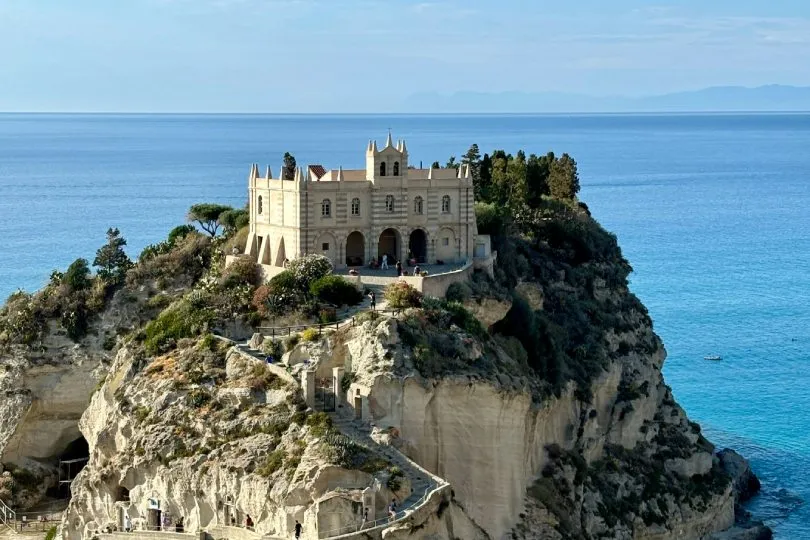 Kirche Santa Maria dell’Isola auf einem felsigen Felsen über dem Meer in Tropea, Kalabrien, mit Treppenwegen am Felsen, Strand und Palmen darunter sowie weitem Blick über das blaue Mittelmeer.