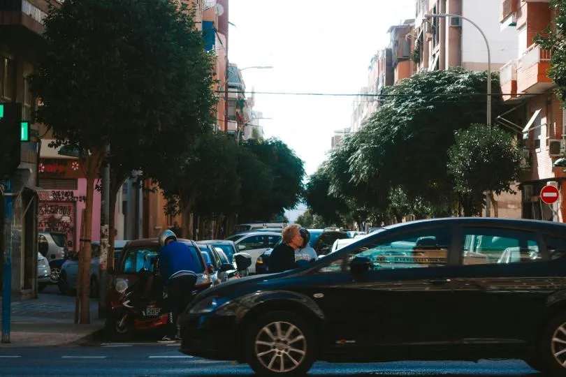 Belebte Stadtstraße mit parkenden Autos, einem vorbeifahrenden schwarzen Wagen, Bäumen am Straßenrand und Fußgängern zwischen den Fahrzeugen.