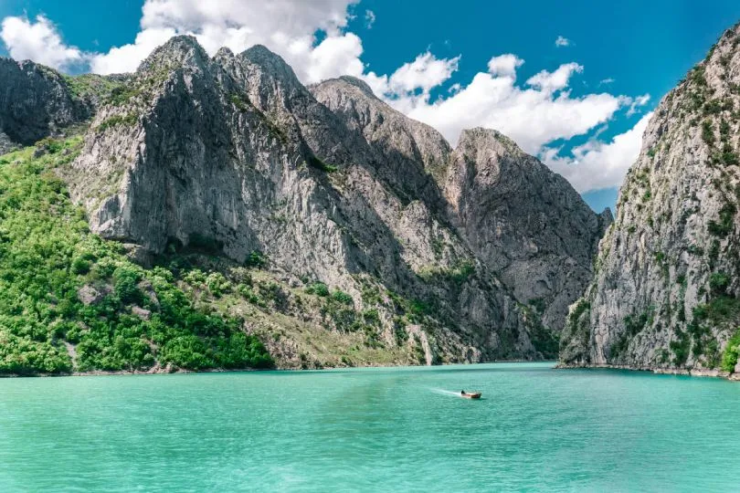 Türkisfarbenes Wasser liegt ruhig zwischen steil aufragenden, felsigen Bergen mit grüner Vegetation. In der Mitte des Sees treibt ein kleines Boot, während darüber ein blauer Himmel mit weißen Wolken zu sehen ist.