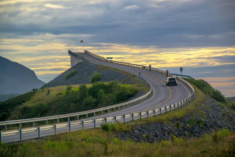 Geschwungene Straße führt über eine Brücke auf einer Anhöhe, ein Auto fährt in Richtung Horizont unter einem dramatischen, bewölkten Himmel.