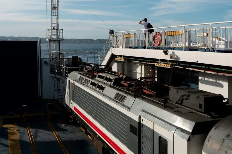 Ein Zug steht auf dem Deck einer Fähre während der Überfahrt zwischen Sizilien und dem italienischen Festland, darüber Passagiere an der Reling mit Blick aufs Meer.