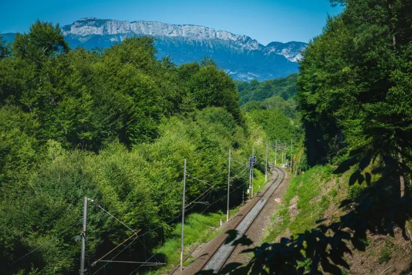 Bahnstrecke führt durch eine grüne, bewaldete Landschaft mit Blick auf Berge im Hintergrund unter blauem Himmel.