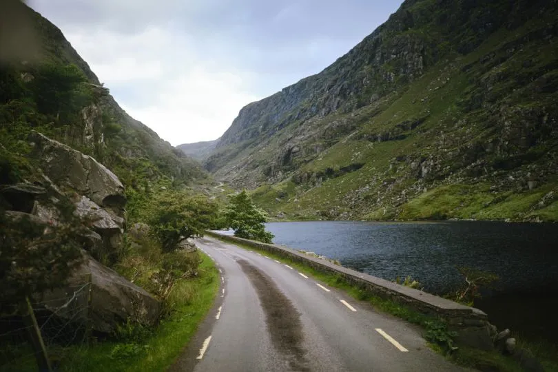 Eine irische Landstraße verkäuft neben einem ruhiges Gewässer, Lichter spiegeln sich auf der Wasseroberfläche.