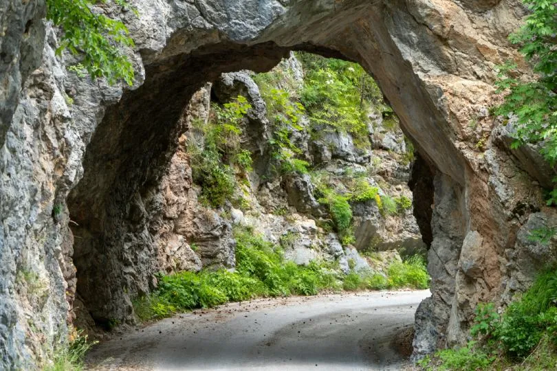 Schmale Straße führt durch einen natürlichen Felsbogen in einer grünen, felsigen Schlucht, umgeben von Bäumen und Vegetation.
