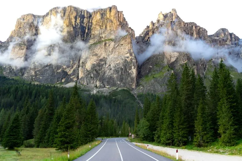 Eine asphaltierte Straße führt durch einen dichten Nadelwald auf hohe, zerklüftete Felsberge zu, deren Gipfel teilweise von Wolken umhüllt sind.