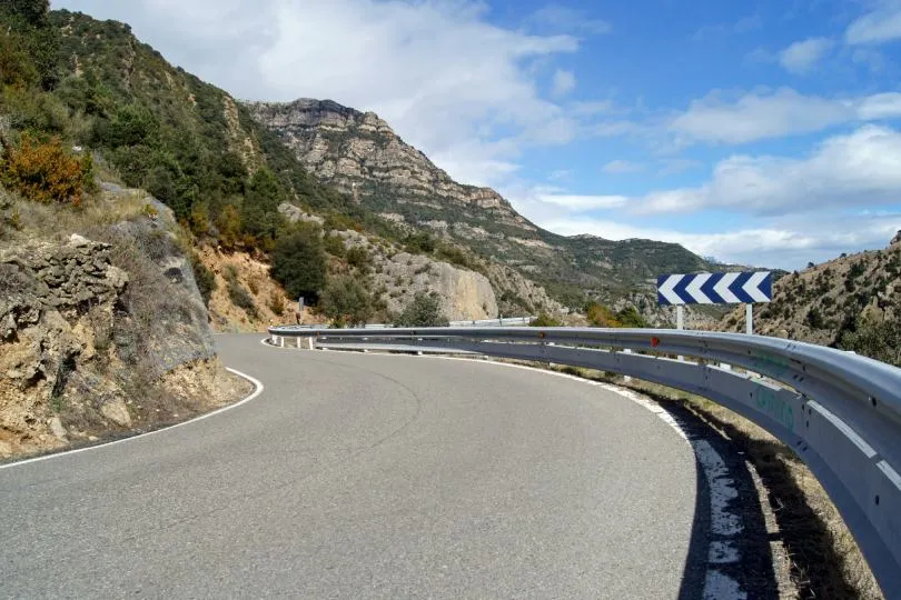 Kurvige Bergstraße mit Leitplanke und Kurvenschild in einer felsigen Landschaft.