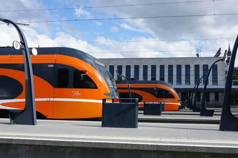 Ein moderner, orangefarbener Personenzug steht an einem Bahnhof mit mehreren Gleisen. Die geschwungenen Bahnsteiglampen und die klare Architektur verleihen der Szene ein futuristisches Erscheinungsbild. Im Hintergrund sind Gebäude sowie wartende Fahrgäste zu erkennen, während der Himmel teils bewölkt ist.