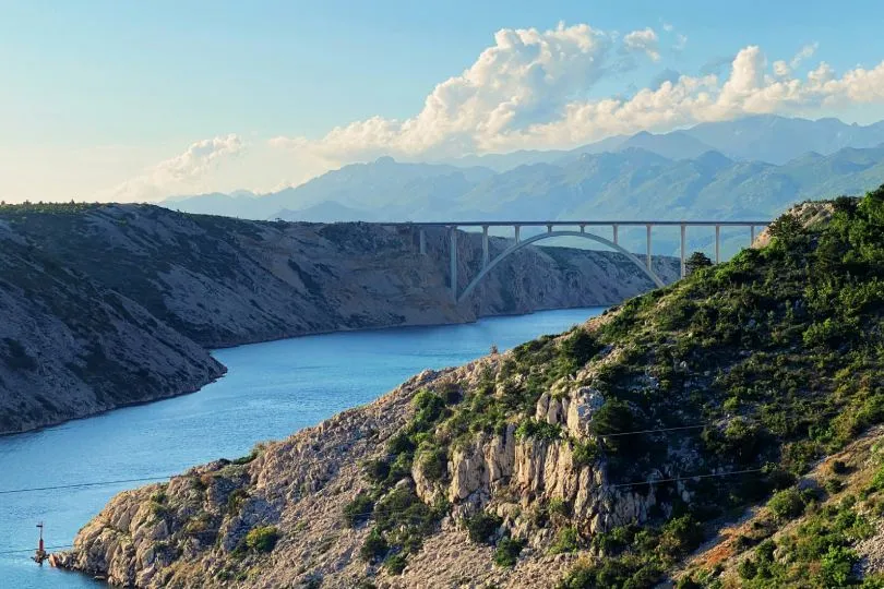 Eine große Bogenbrücke spannt sich über einen tief eingeschnittenen Meeresarm oder Fluss zwischen kargen, felsigen Hügeln. Das Wasser schimmert blau, im Hintergrund erheben sich mehrere Bergketten unter einem Himmel mit lockeren Wolken.