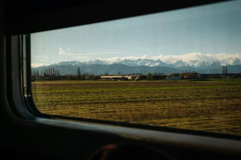 Blick aus einem Zugfenster auf eine vorbeiziehende ländliche Landschaft mit Feldern und kleinen Gebäuden, dahinter eine lange Bergkette mit schneebedeckten Gipfeln unter klarem Himmel.