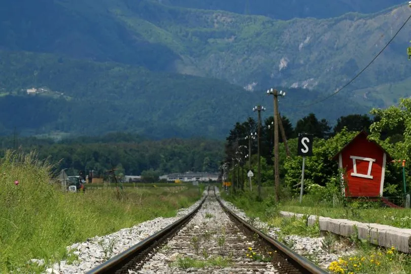 Eisenbahngleise führen durch eine ländliche Landschaft mit Wiesen und kleinen Häusern, während im Hintergrund bewaldete Berge aufragen und neben der Strecke ein kleines rotes Bahnhäuschen steht.