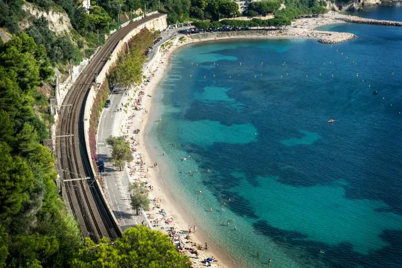 Luftaufnahme einer türkisfarbenen Bucht mit Sandstrand, an dem viele Menschen baden und liegen, während direkt daneben eine Eisenbahnstrecke entlang der felsigen Küste durch grüne Hügel verläuft.
