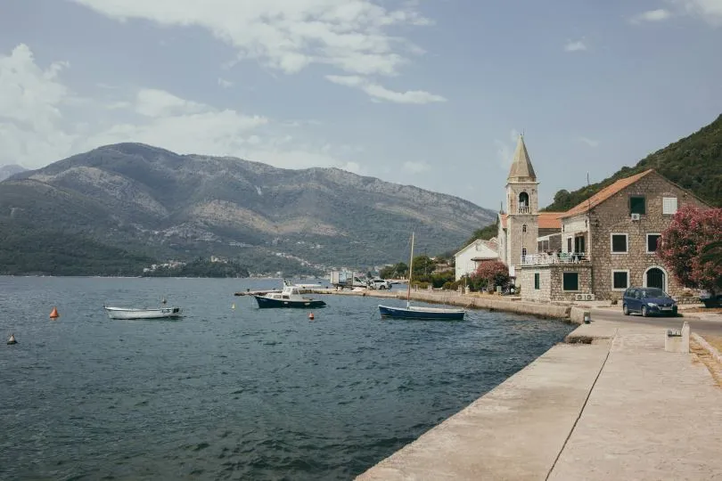 Kleiner Küstenort mit Kirche und Steinhäusern am Wasser, mehrere Boote liegen im ruhigen Hafen vor einer bergigen Landschaft.