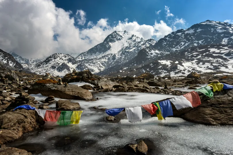 Himalaya-Landschaft im Mustang-Gebirge in Nepal mit schneebedeckten Gipfeln und bunten Gebetsfahnen über einem vereisten Bergbach.