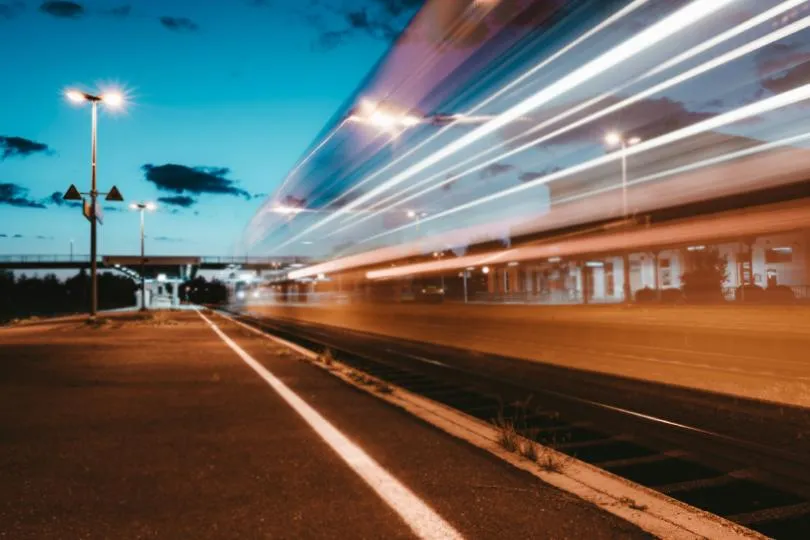 Zug fährt mit Bewegungsunschärfe durch einen beleuchteten Bahnhof bei Dämmerung, Bahnsteig und Gleise im Vordergrund.