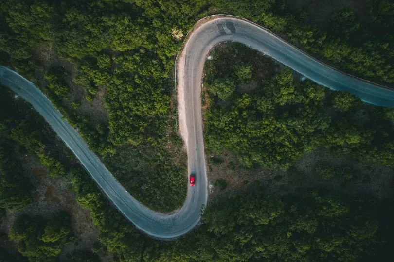 Luftaufnahme einer kurvenreichen Straße in S-Form, die durch dichten Wald führt, mit einem einzelnen roten Auto auf der Strecke.