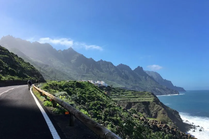Küstenstraße mit Wanderern, Blick auf grüne Terrassen, steile Berge und das Meer unter blauem Himmel.