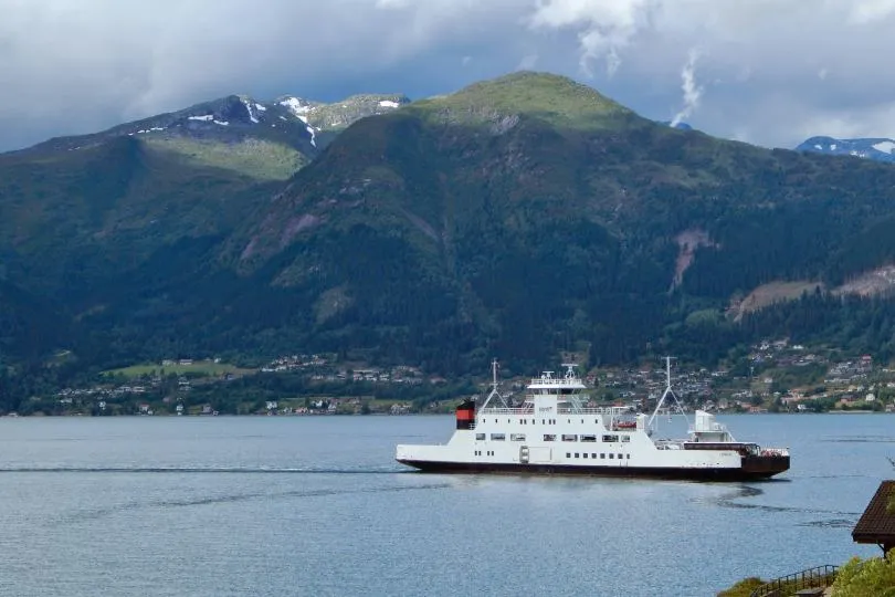 Fähre fährt über einen ruhigen Fjord, im Hintergrund liegen grüne Berge und eine kleine Siedlung am Ufer.