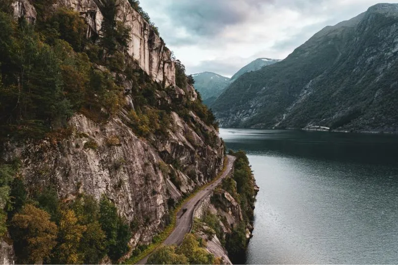 Schmale Straße verläuft entlang einer steilen Felswand am Ufer eines ruhigen Sees, umgeben von bewaldeten Bergen unter bewölktem Himmel.