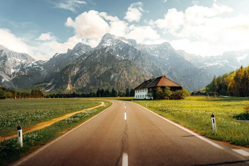 Eine Landstraße führt durch eine weite Wiese zu einem einzelnen Haus vor einer beeindruckenden Bergkulisse mit schneebedeckten Gipfeln und teilweise bewölktem Himmel.