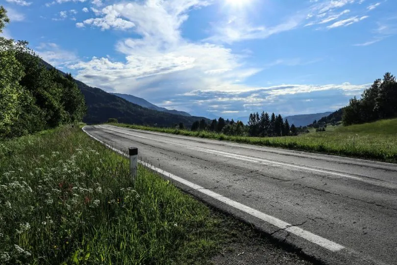 Landstraße verläuft durch eine hügelige, grüne Landschaft mit Wiesen und Bäumen, im Hintergrund sanfte Berge unter einem weiten Himmel mit Wolken.