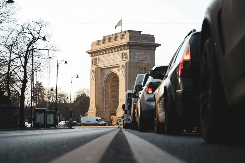 Eine Reihe von Autos steht im Stadtverkehr vor einem monumentalen Triumphbogen. Die Aufnahme ist aus niedriger Perspektive von der Straße aus gemacht, wodurch die Fahrzeuge im Vordergrund und das historische Bauwerk im Hintergrund besonders betont werden.
